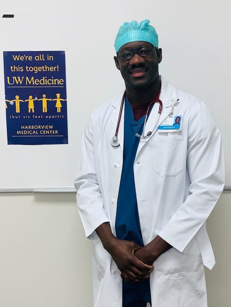 Ebou M A Cham stands in clinical attire in front of a sign that says, "We're all in this together. UW Medicine. Harborview Medical Center."