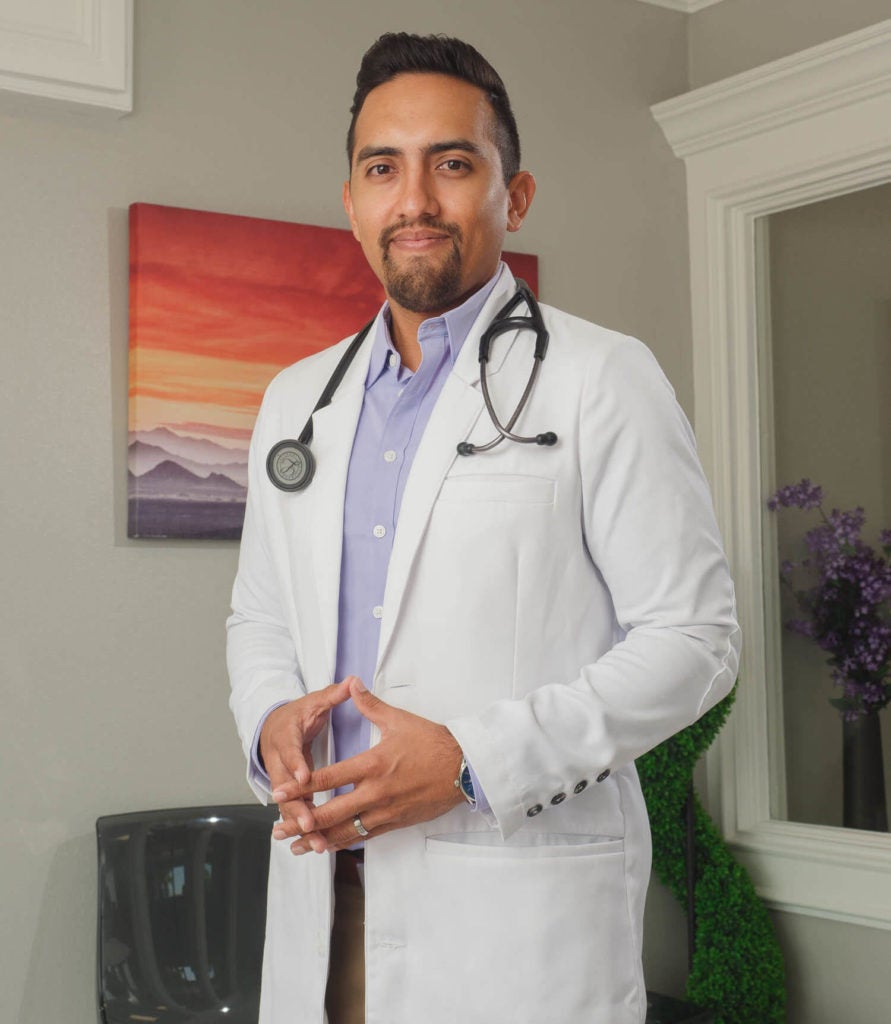 Michael Menchaca, in a white coat and stethoscope, stands in his clinic.