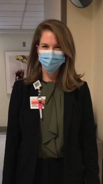 Maggie Raymond stands in business attire and a surgical mask in a hospital hallway.