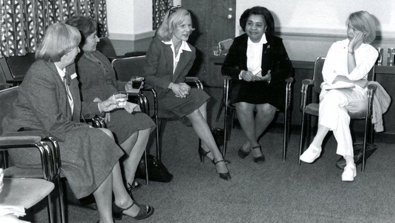 In a historical black and white photo, five women sit together at an event in a conference room.