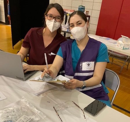 Jessica Landau and Natalie Jarian, left to right, pose in clinical attire at a COVID-19 vaccine site.