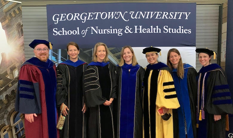 At Commencement 2021 at Nationals Park are, left to right, Dr. Matthew Kavangh, Dr. Myriam Vuckovic, Dr. Gresenz, Dr. Jan LaRocque, Dr. Roxanne Mirabal-Beltran, Dr. Blythe Shepard, and Dr. Elizabeth Sloss pose in academic gowns in front of sign and photo that reads Georgetown University School of Nursing & Health Studies
