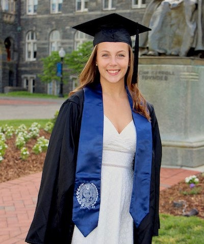 Karly Ward in her graduation gown and a graduation stole with the Georgetown seal stands in front of the John Carroll statue on Georgetown's campus.