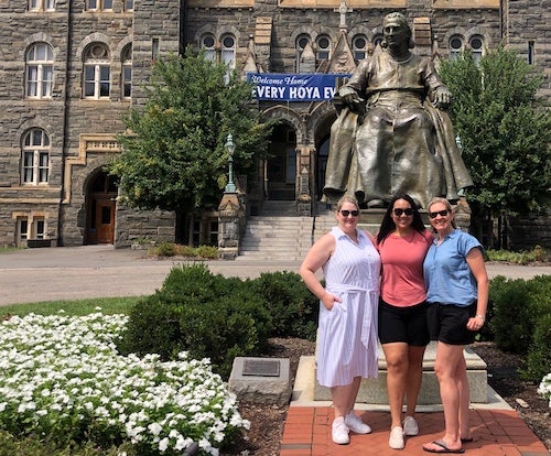 Three graduates stand in front of the John Carroll statue in front of Healy Hall on Georgetown's campus