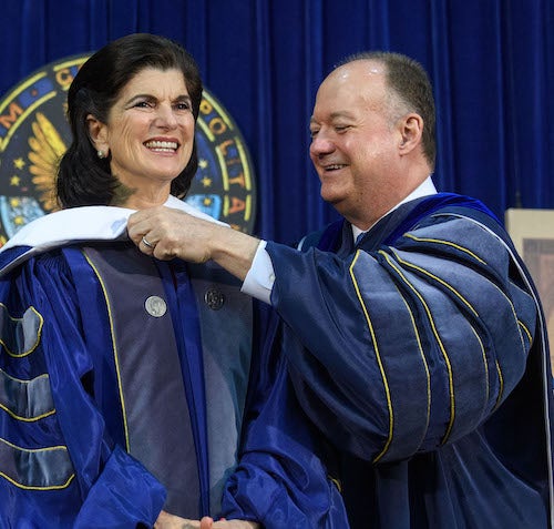 Georgetown President John J. DeGioia helps Luci Baines Johnson with her academic hood during Commencement with a blue curtain and a seal of Georgetown University behind them.