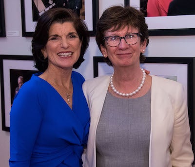 Luci Baines Johnson and Patricia Cloonan pose together in front of framed photographs on the wall.