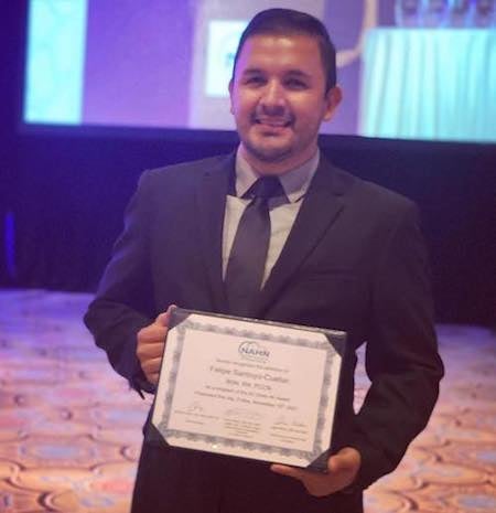 Felipe Santoyo-Cuellar holds his award certificate in an indoor setting with a movie screen behind him