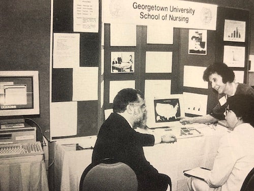 Dr. Virginia Saba, in front of a 1980s computer terminal and display area, describes her classification system to two people who are seated at the table.