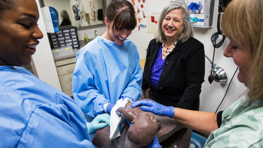 Students use the nursing birthing simulator as professors look on