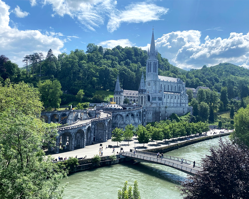 A view of the Sanctuary of Our Lady of Lourdes, a towering cathedral next to a green river framed by rolling tree-covered hills