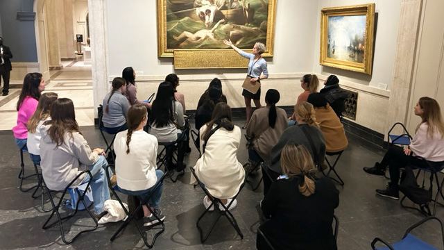 A woman gestures to a painting on the wall at the National Gallery of Art while nursing students sitting on portable chairs listen to what she is saying