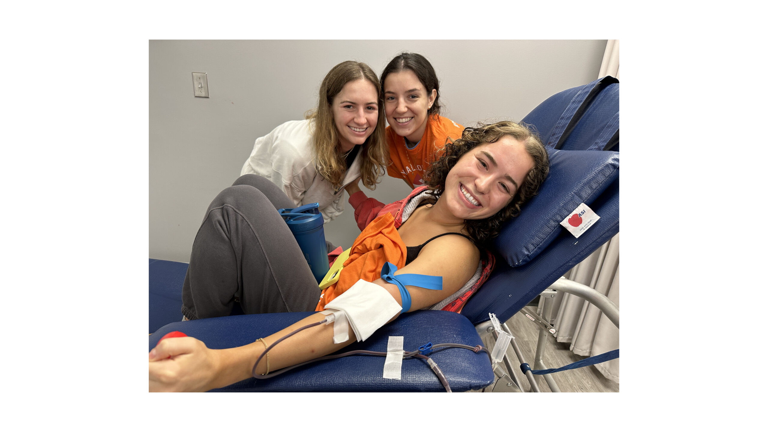 Three students smiling at a blood drive while one is donating blood.