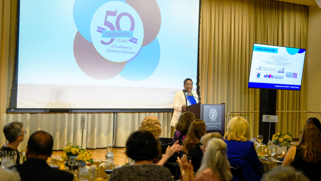Dean Roberta Waite speaking at a podium with a large screen behind her displaying a logo with the words Celebrating 50 Years of Excellence in Midwifery Education