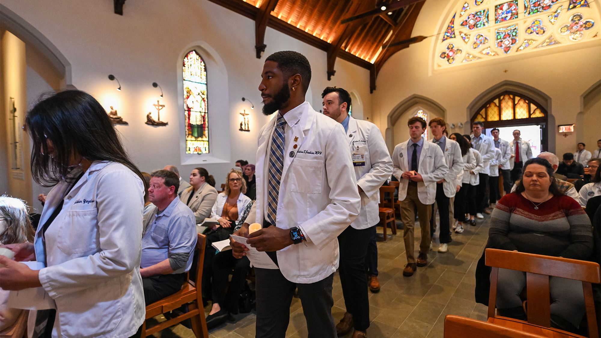 Students in white coats file into Dahlgren Chapel while congregants sit in pews around them