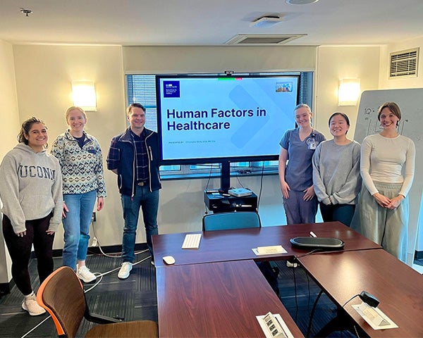 A group of individuals stands near a screen at the front of the room on which is projected the words Human Factors in Healthcare