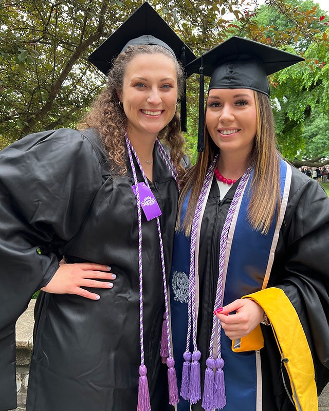 Kayla and Jordan stand side by side dressed in their academic regalia