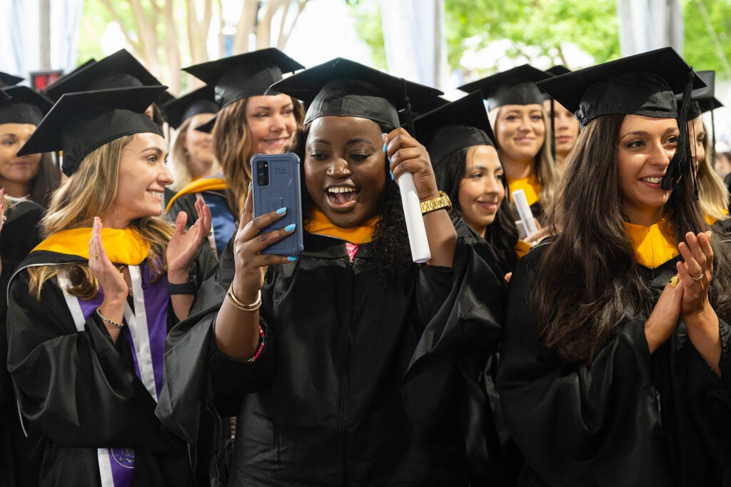A graduate looks into her phone and raises an arm in celebration while other students crowd around her