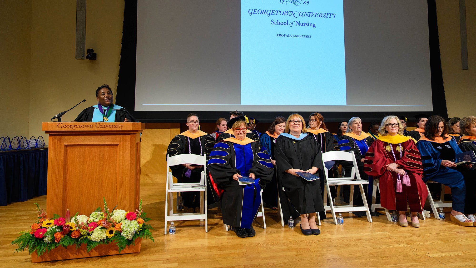 Dean Waite speaks from a podium at Tropaia while the nursing faculty sit in chairs onstage
