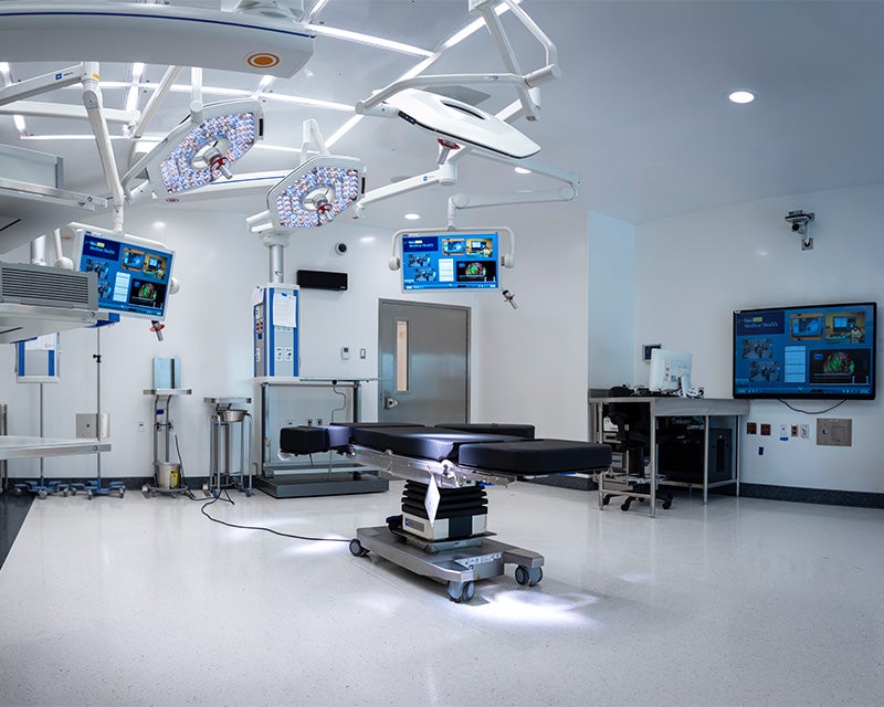 An operating room featuring a table in the middle of a white room with screens on the wall and overhead
