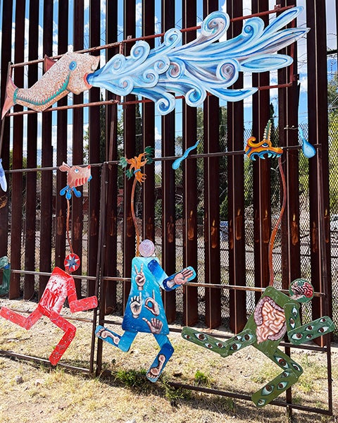 The border wall on the Mexican side is vertical iron bars overlaid with a sculpture depicting people running and a fish exhaling a gush of water