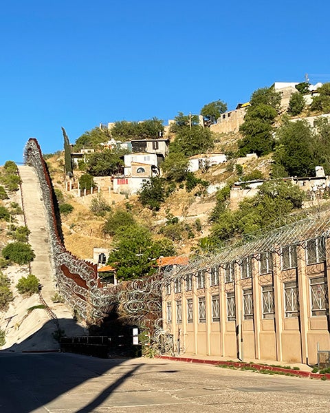 A dry hillside with a fence topped with spirals of razor wire divides buildings on the U.S. Mexico border