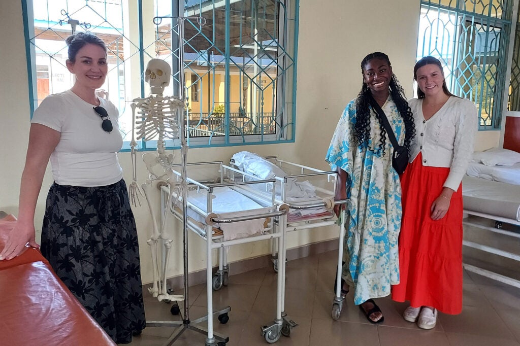 The three students stand amid beds and bassinets in a hospital setting with a model skeleton behind them