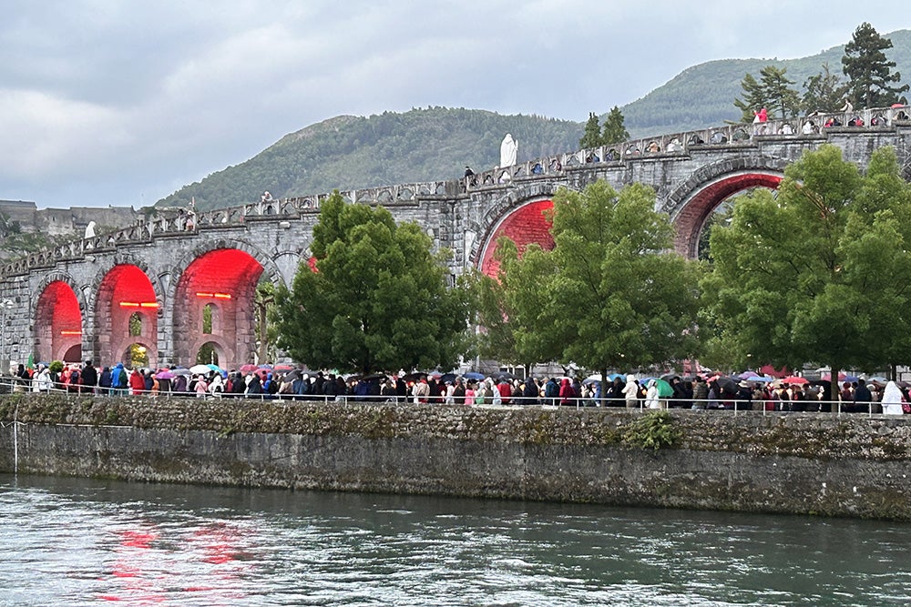 A line of pilgrims runs along the shoreline of the river below the cathedral of Lourdes