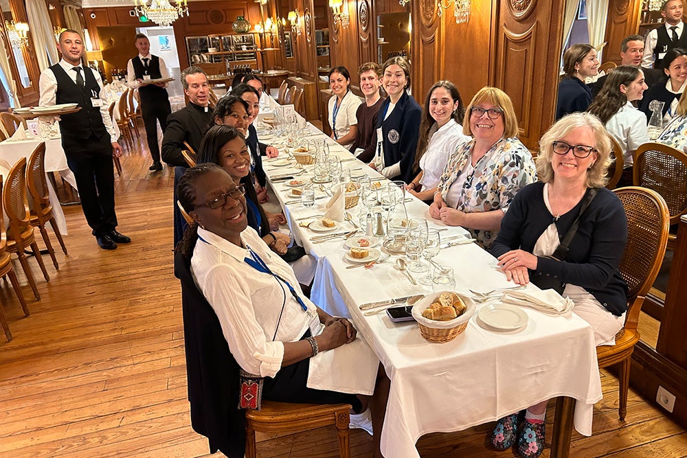 A group of people sit at a long table that is covered in a white tablecloth with waiters standing nearby with trays