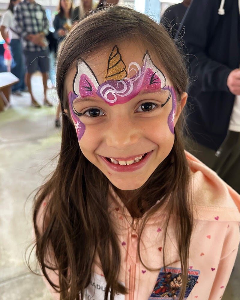 A young girl with unicorn ears and horn painted on her forehead smiles broadly