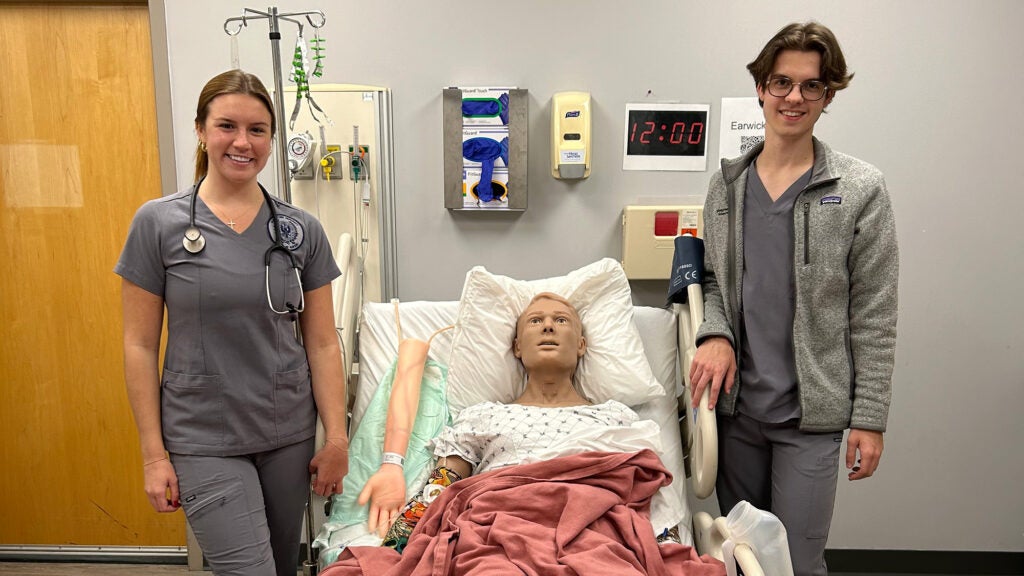 Two nursing students stand next to a bed where a simulation dummy is lying