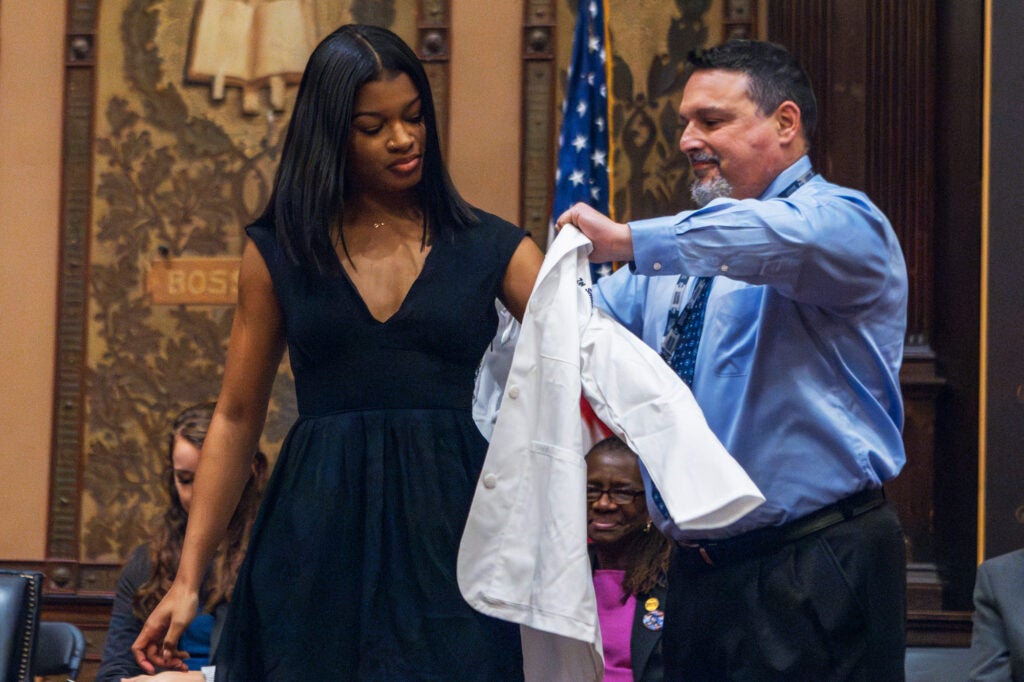 A nursing school faculty member helps student Chloe into her white coat on the stage in Gaston Hall