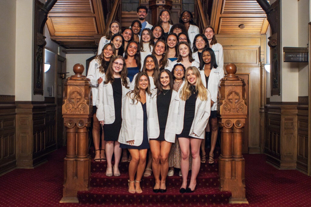 A group of 23 nursing students all wearing their white coats stands together on a staircase in Healy Hall