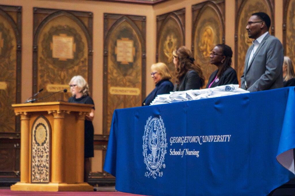 Sarah Vittone speaks at a podium while the dean and other faculty stand onstage behind her in Gaston Hall