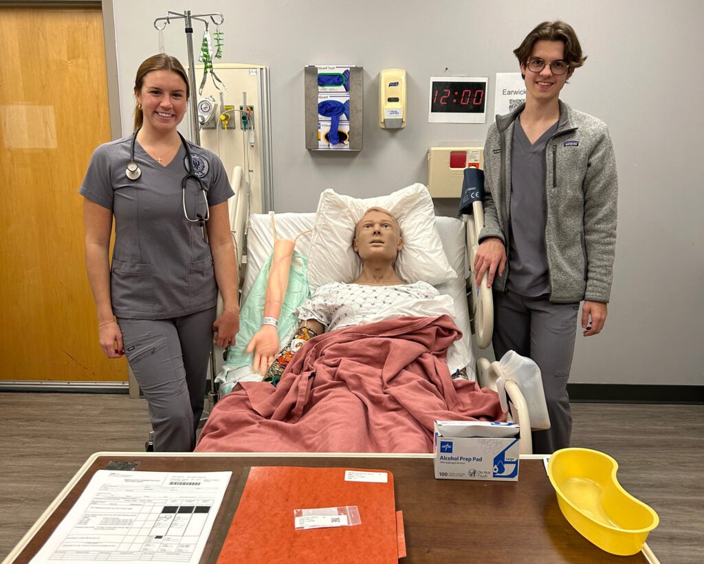 Two students stand on either side of a hospital bed in which a simulator dummy lies in the GUSON Sim Lab