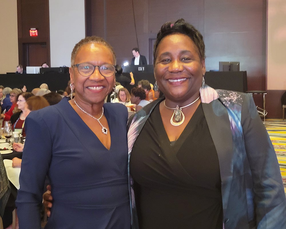 Jemmott and Waite stand together in a large room, people are seated at tables behind them