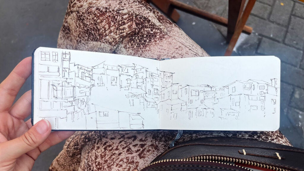 A closeup of a hand holding an open sketchbook on which there is a line drawing of houses clustered together
