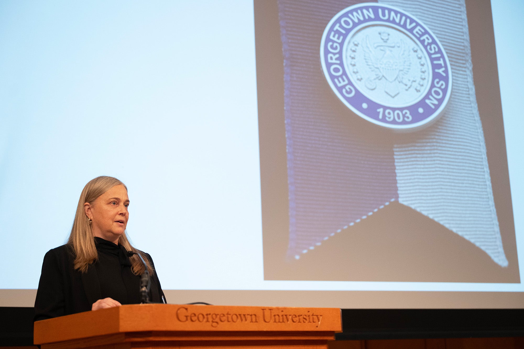 Kelli Giffin speaks from a podium, behind her is a projected image of a School of Nursing pin