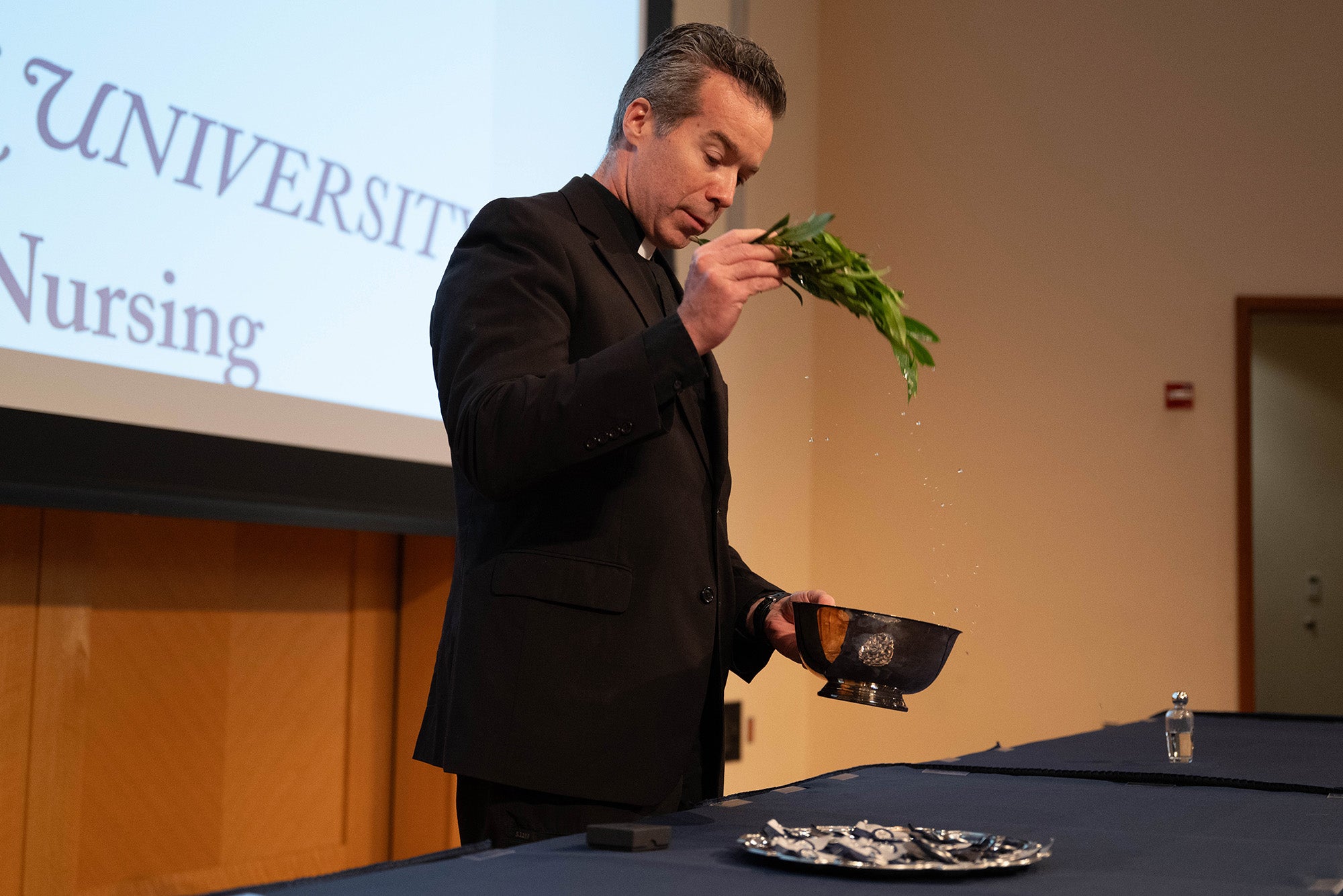 A priest uses a green leafy branch to sprinkle water onto pins on a silver platter sitting on a table in front of him
