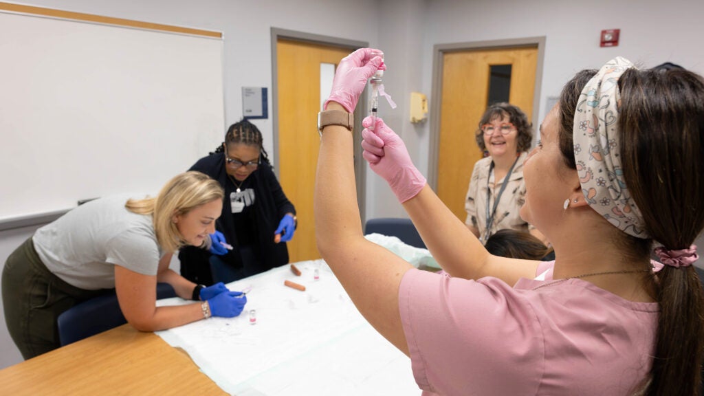 A nursing student loads a syringe while in the background two students practice injections using a hotdog