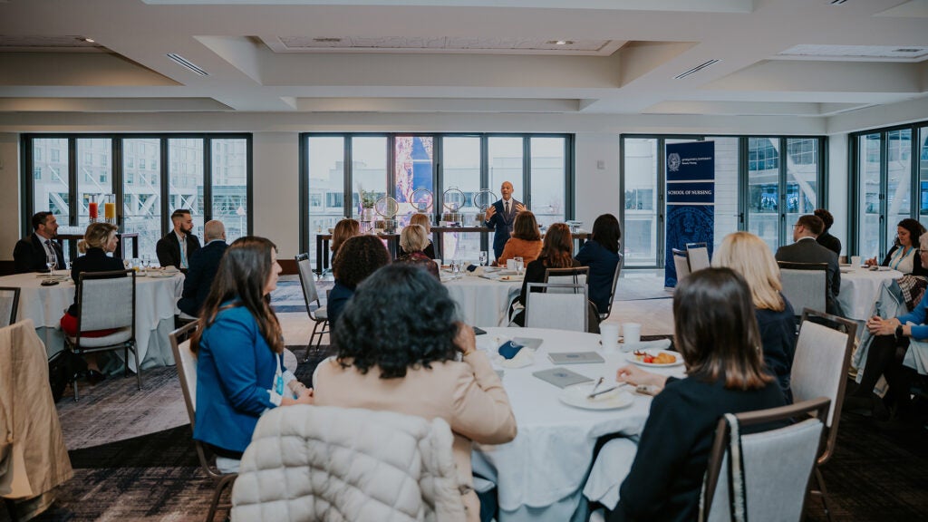 Stephan Davis speaks before people seated at round tables in a conference room lines with windows