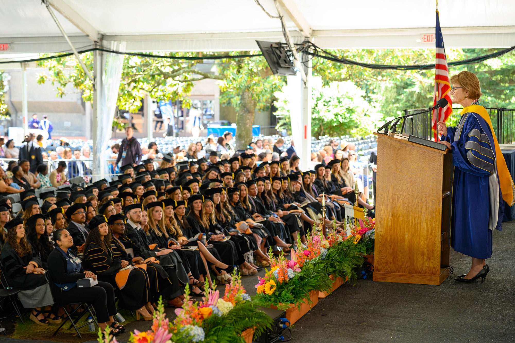 Students in caps and gowns sit in rows of chairs in front of Mary Wakefield speaking from a podium on a stage