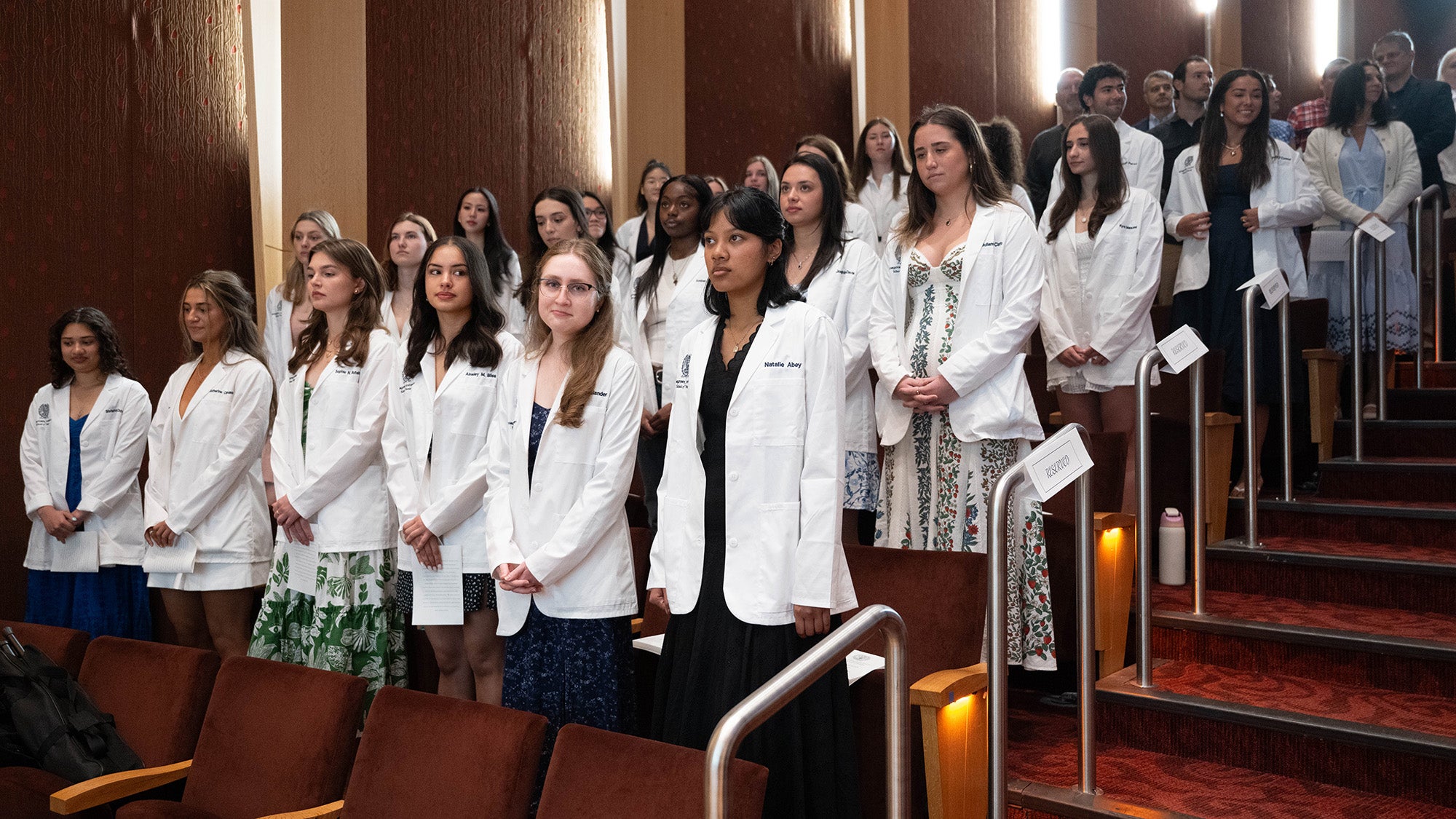 Nursing students in white coats stand in an auditorium