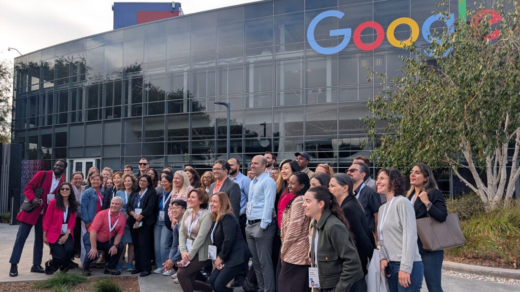A large group of people stand before a glass fronted building with a sign reading Google on it