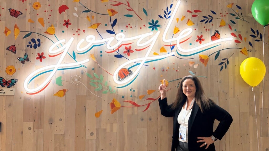 Ogg-Gress points to a neon script sign on a wall that reads Google surrounded by painted flowers and butterflies, two balloons are nearby