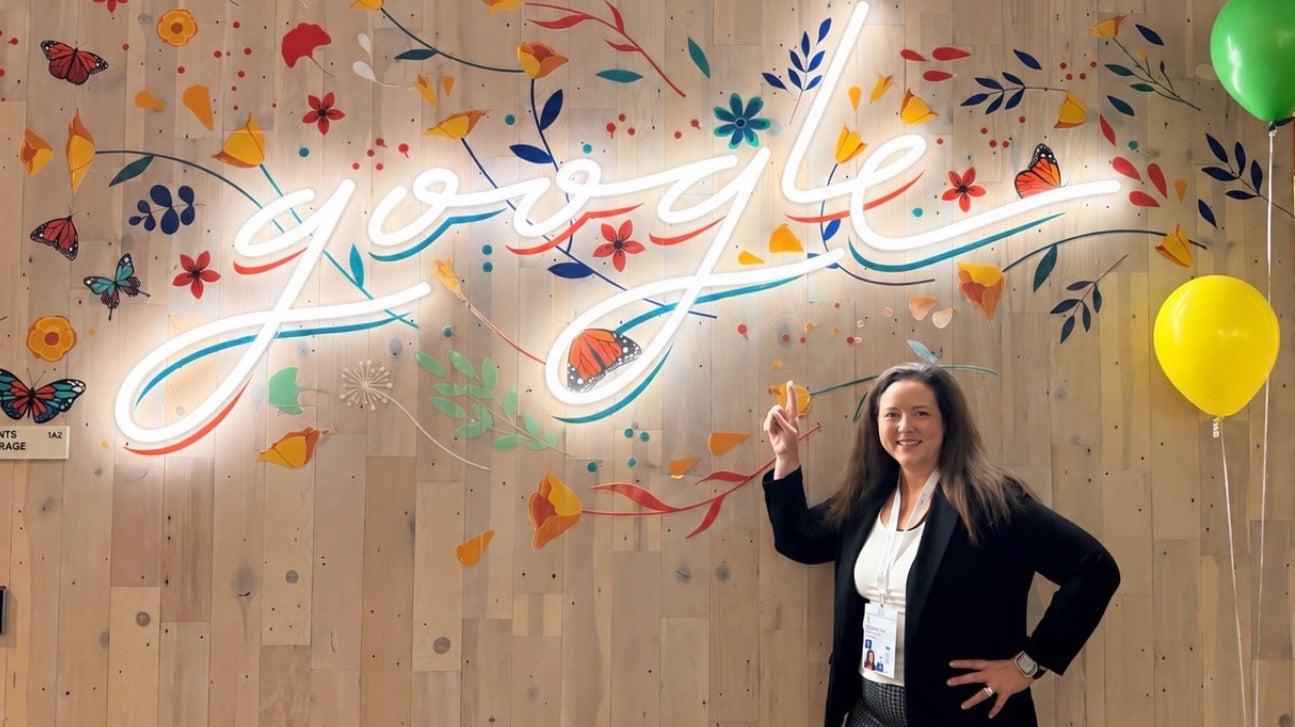 Ogg-Gress points to a neon script sign on a wall that reads Google surrounded by painted flowers and butterflies, two balloons are nearby